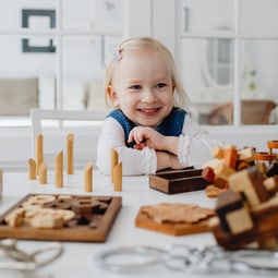 Lächelndes Kleinkind mit blonden Haaren, das an einem Tisch mit Holzspielzeugen spielt